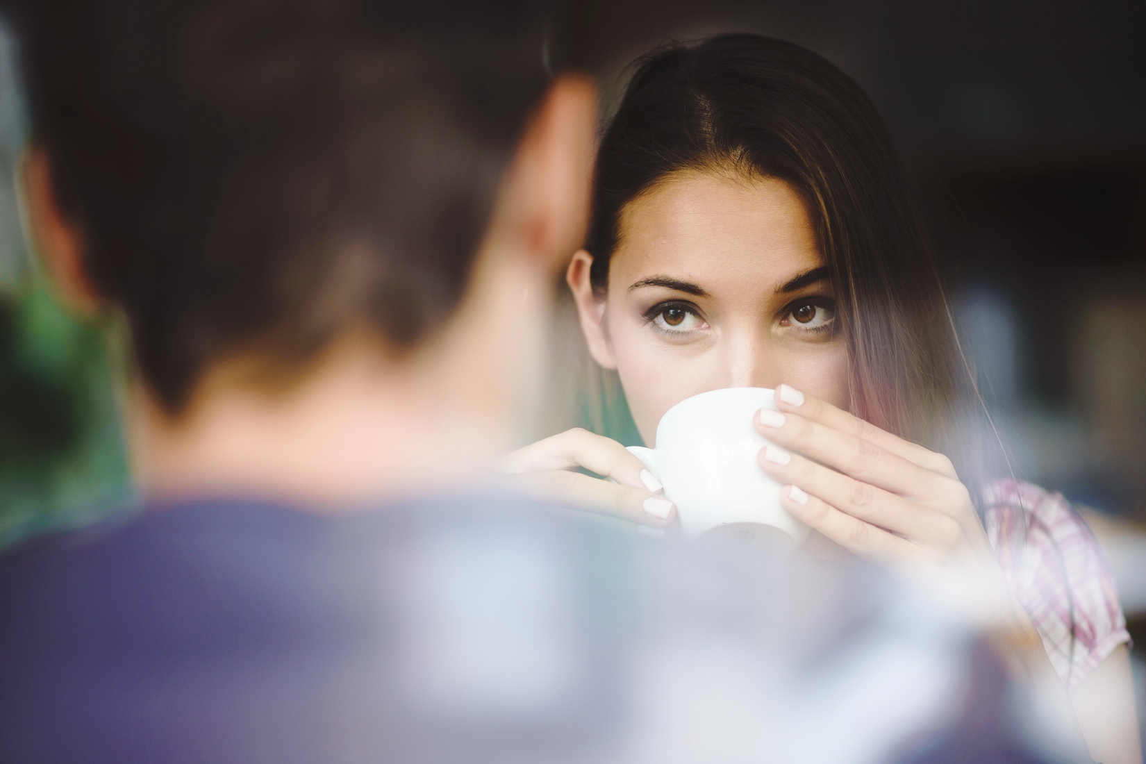 A woman with long brown hair looks over the rim of a white cup she is holding, appearing thoughtful. The blurred silhouette of another person is in the foreground, suggesting a conversation or shared moment.