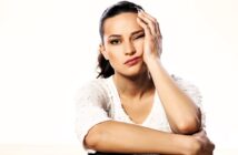 A woman with dark hair in a ponytail rests her head on her hand, winking with one eye. She's wearing a white shirt and appears to be in a thoughtful or playful mood against a plain light background.