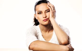 A woman with dark hair in a ponytail rests her head on her hand, winking with one eye. She's wearing a white shirt and appears to be in a thoughtful or playful mood against a plain light background.