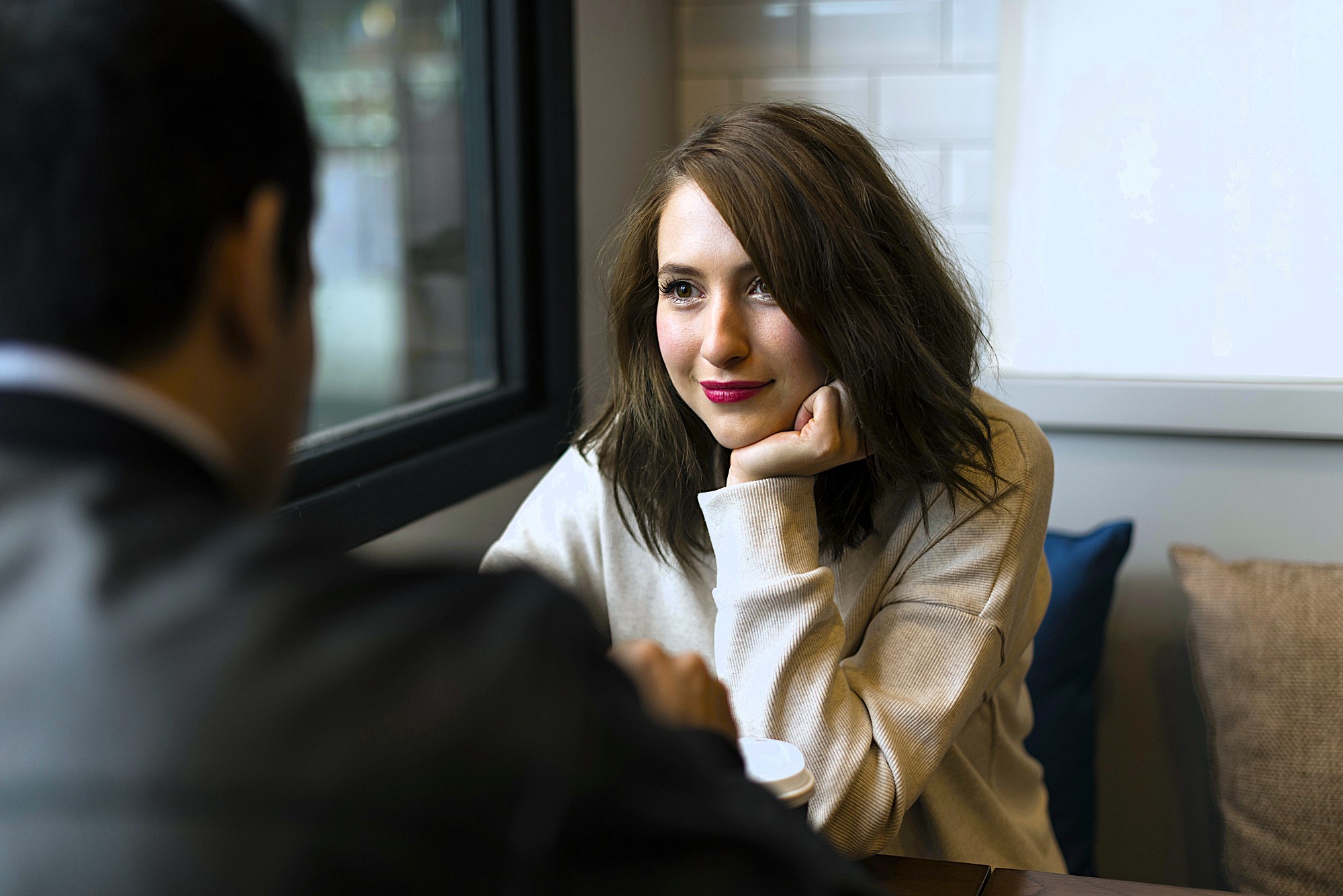 A woman with shoulder-length brown hair and a beige sweater smiles while resting her chin on her hand, looking at a man sitting across from her. They are seated indoors with a window in the background.