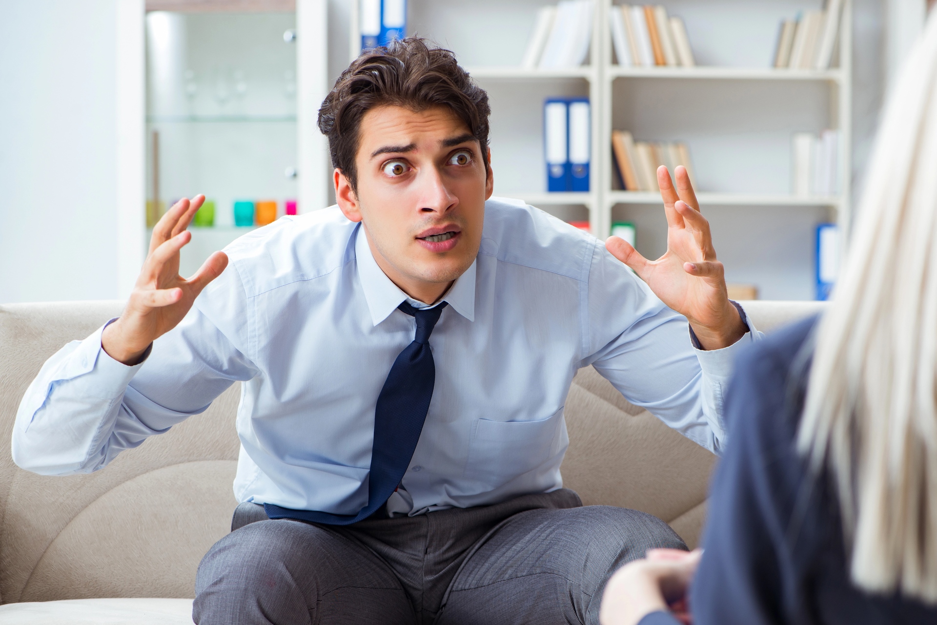 A man in a blue shirt and tie sits on a couch with his arms raised, expressing frustration during a conversation. Shelves with books and binders are in the background, and a woman is partially visible, sitting opposite him.