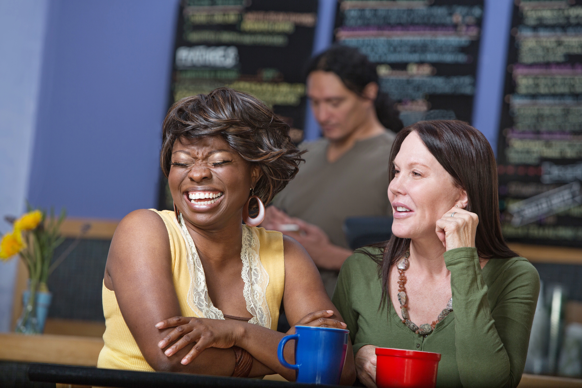 Two women sitting at a café table, both smiling and holding coffee mugs. The woman on the left is laughing, and the woman on the right is chatting. A blurred figure stands in the background near a menu board on the wall.