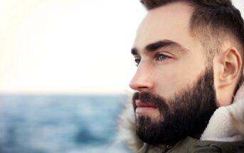 A bearded man gazes thoughtfully into the distance, standing near the ocean. He wears a jacket with a furry collar. The background shows a blurred seascape under a clear sky.