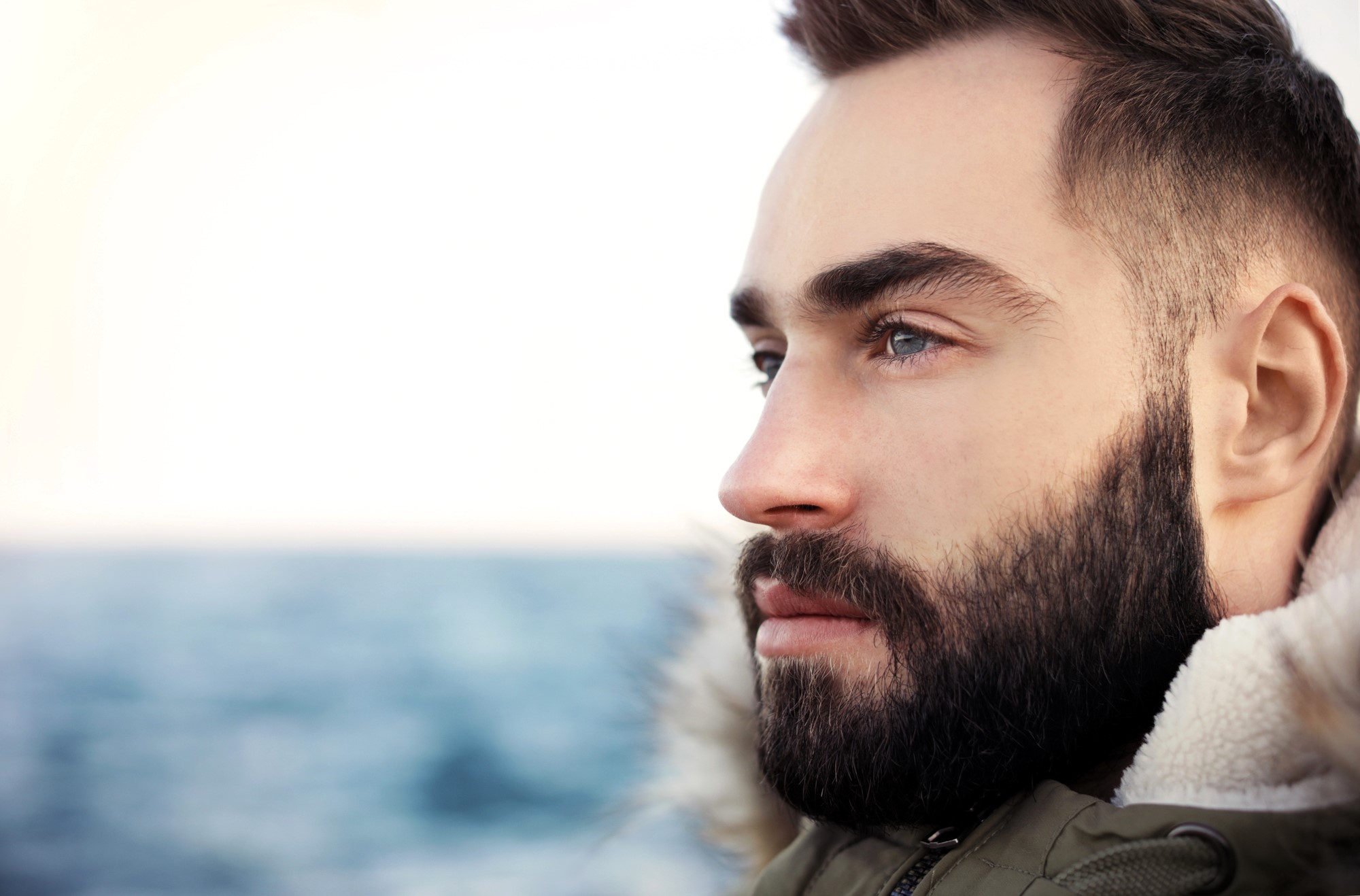 A bearded man gazes thoughtfully into the distance, standing near the ocean. He wears a jacket with a furry collar. The background shows a blurred seascape under a clear sky.