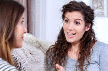 Two women are sitting on a couch engaged in a conversation. The woman on the right has curly hair and is smiling while talking. The woman on the left is listening attentively. A crocheted pillow is visible behind them.