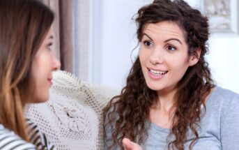 Two women are sitting on a couch engaged in a conversation. The woman on the right has curly hair and is smiling while talking. The woman on the left is listening attentively. A crocheted pillow is visible behind them.