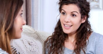 Two women are sitting on a couch engaged in a conversation. The woman on the right has curly hair and is smiling while talking. The woman on the left is listening attentively. A crocheted pillow is visible behind them.