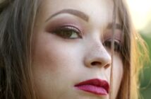 Close-up of a person's face with a serious expression. They have long hair, pink eye makeup with eyeliner, and prominent dark red lipstick. The lighting highlights their features, with a blurred green background.