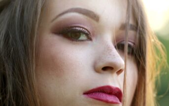 Close-up of a person's face with a serious expression. They have long hair, pink eye makeup with eyeliner, and prominent dark red lipstick. The lighting highlights their features, with a blurred green background.