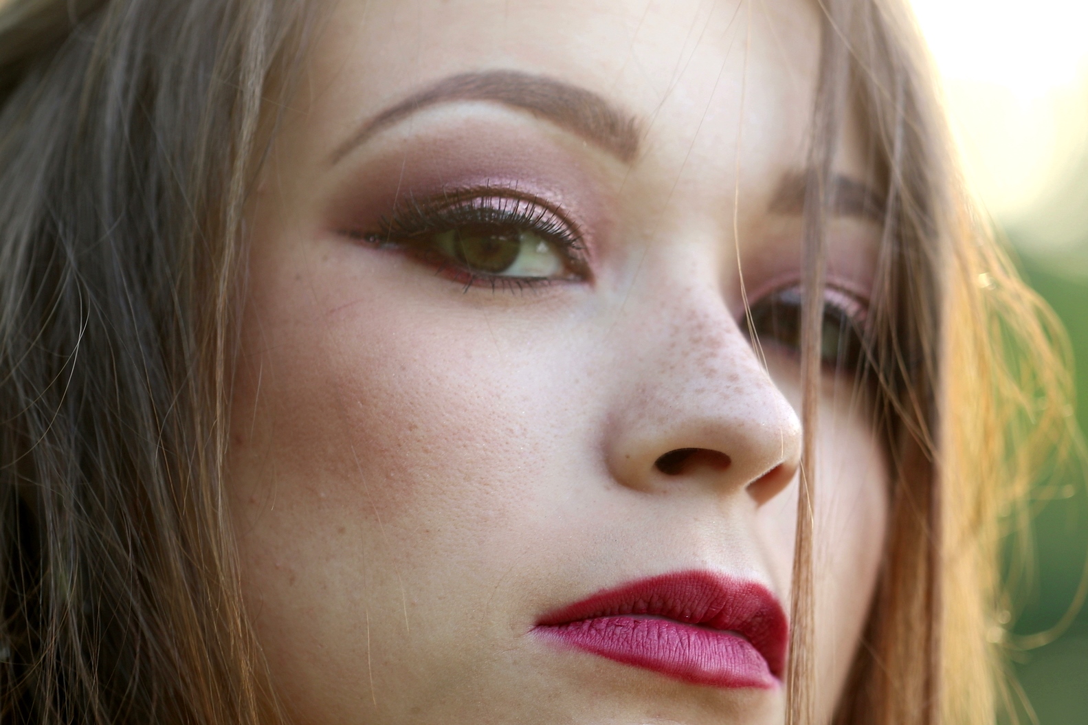 Close-up of a person's face with a serious expression. They have long hair, pink eye makeup with eyeliner, and prominent dark red lipstick. The lighting highlights their features, with a blurred green background.