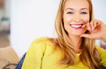 A woman with long blonde hair smiles at the camera. She is wearing a bright yellow top and is sitting on a beige sofa. The background is softly blurred, creating a warm and inviting atmosphere.