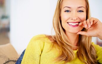 A woman with long blonde hair smiles at the camera. She is wearing a bright yellow top and is sitting on a beige sofa. The background is softly blurred, creating a warm and inviting atmosphere.