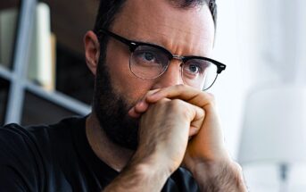 A man with glasses and a beard is gazing intently, chin resting on his hands. He appears deep in thought, with a bookshelf and a lamp in the blurred background.