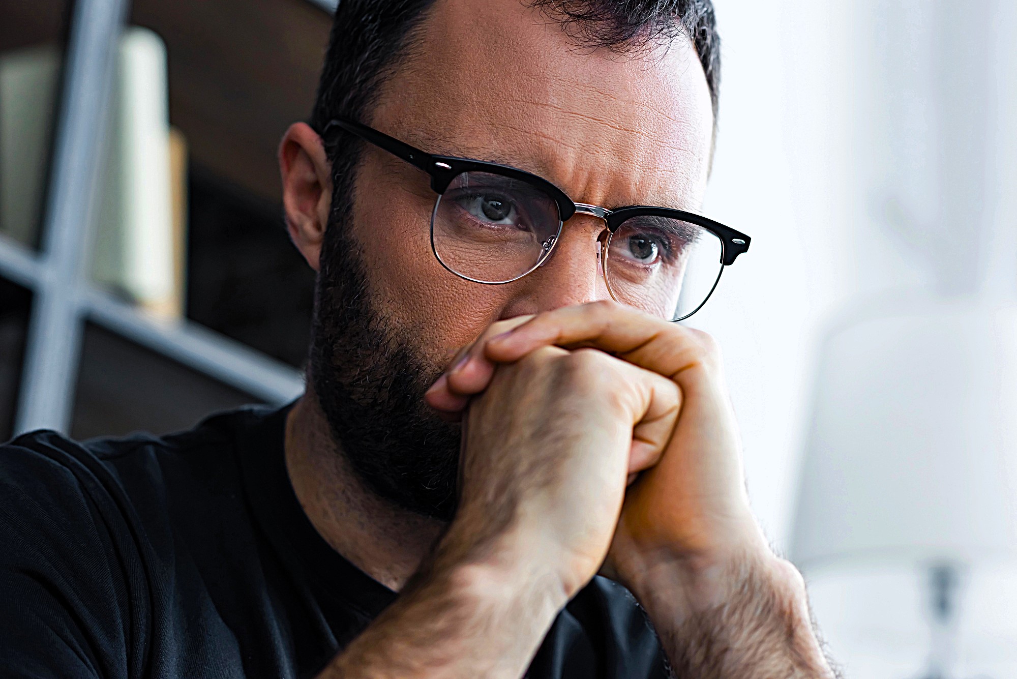 A man with glasses and a beard is gazing intently, chin resting on his hands. He appears deep in thought, with a bookshelf and a lamp in the blurred background.