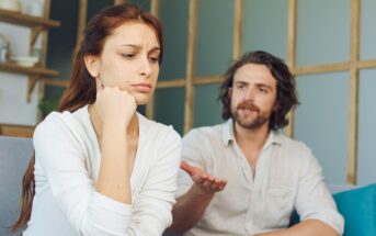 A woman sits pensively on a couch, resting her chin on her hand, appearing upset. A man sits nearby, gesturing with one hand, seemingly talking to her. The room is softly lit, with shelves and decor in the background.