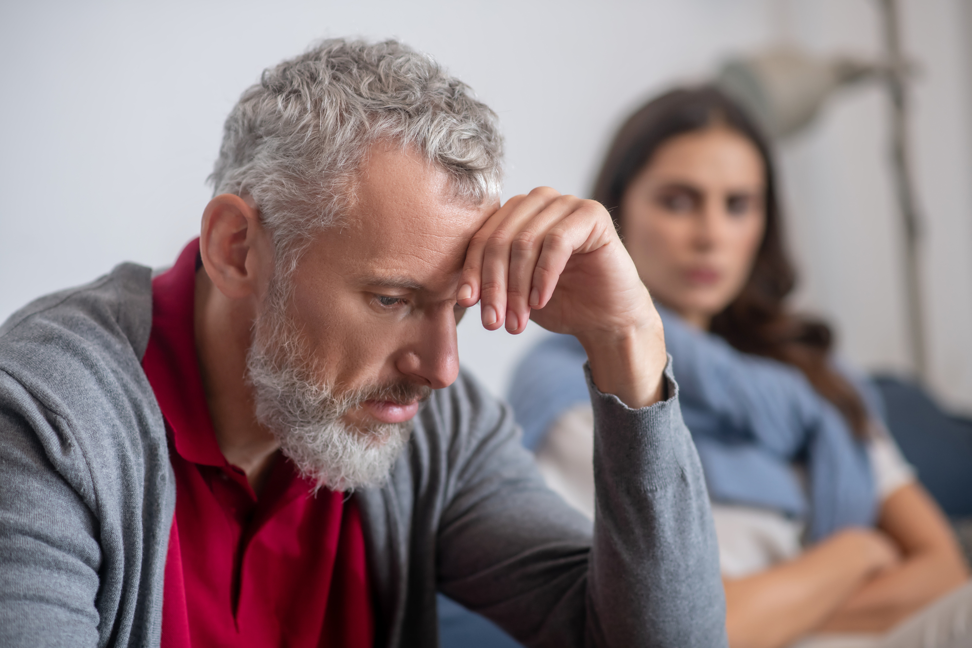 A man with gray hair and a beard looks stressed, resting his head on his hand. In the blurred background, a woman with long brown hair appears concerned, sitting with her arms crossed. They are indoors on a couch.