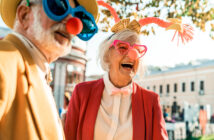 Two elderly individuals are enjoying a festive outdoor event. Both are wearing large, colorful sunglasses, clown noses, and playful costumes. The woman is laughing heartily, wearing red heart-shaped glasses and a bow headband. The man is in a yellow hat.