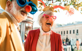 Two elderly individuals are enjoying a festive outdoor event. Both are wearing large, colorful sunglasses, clown noses, and playful costumes. The woman is laughing heartily, wearing red heart-shaped glasses and a bow headband. The man is in a yellow hat.