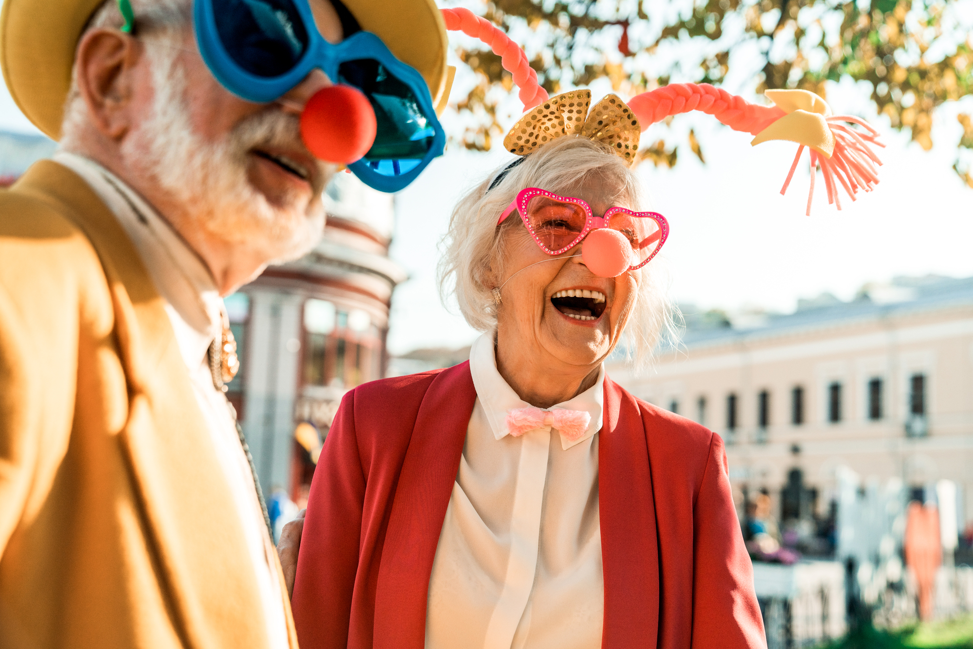 Two elderly individuals are enjoying a festive outdoor event. Both are wearing large, colorful sunglasses, clown noses, and playful costumes. The woman is laughing heartily, wearing red heart-shaped glasses and a bow headband. The man is in a yellow hat.