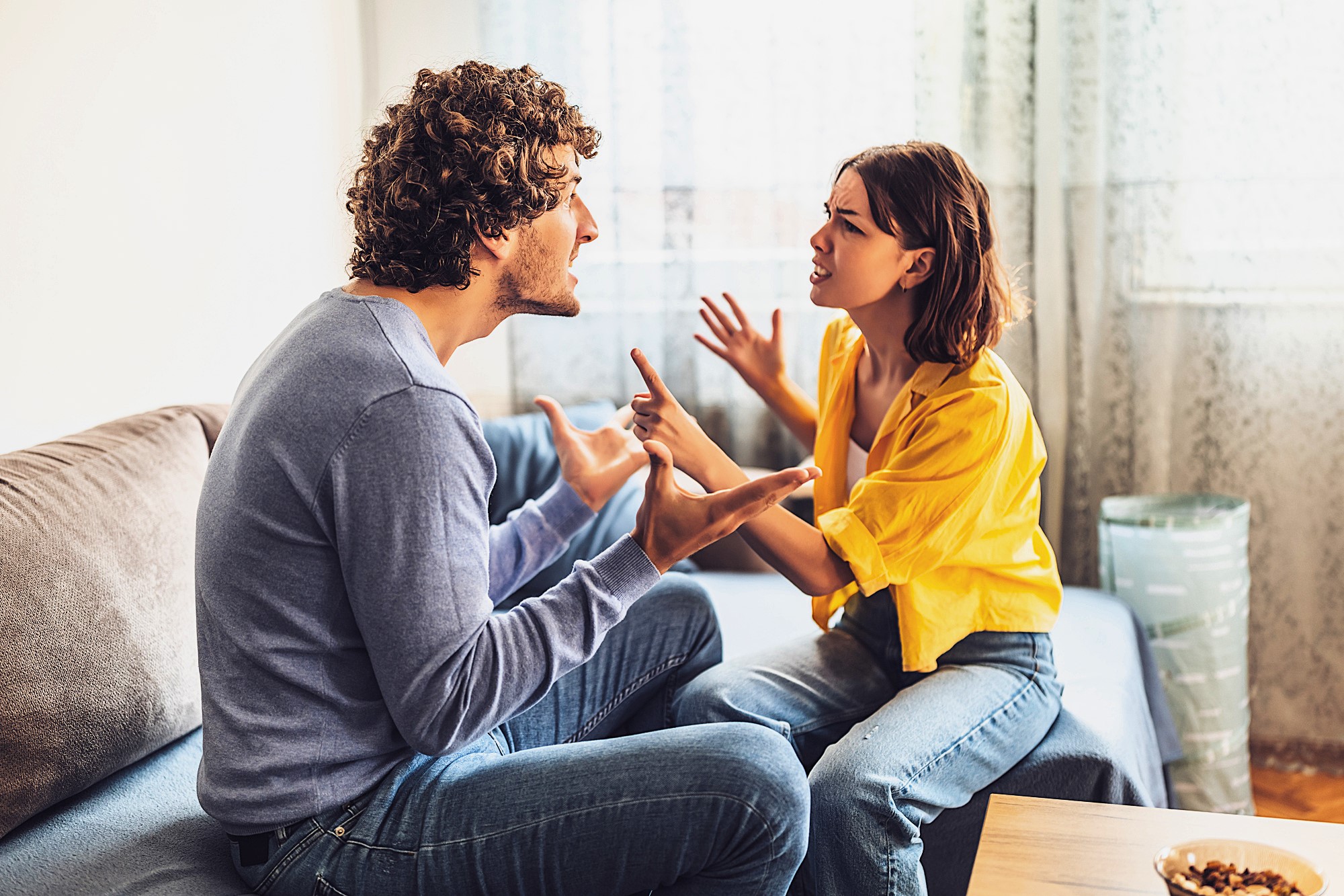 A man and woman are sitting on a sofa, engaged in an intense argument. The man, in a light blue sweater, gestures with open hands, while the woman, wearing a yellow blouse, responds emphatically with raised hands. They are in a living room setting.