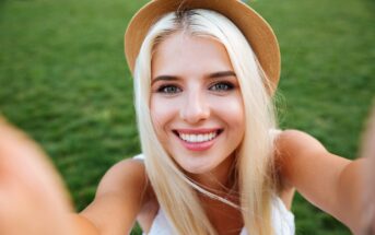 A person with long blonde hair wearing a straw hat takes a selfie outdoors. They are smiling widely, with a grassy background. The sun is shining, creating a bright and warm atmosphere.