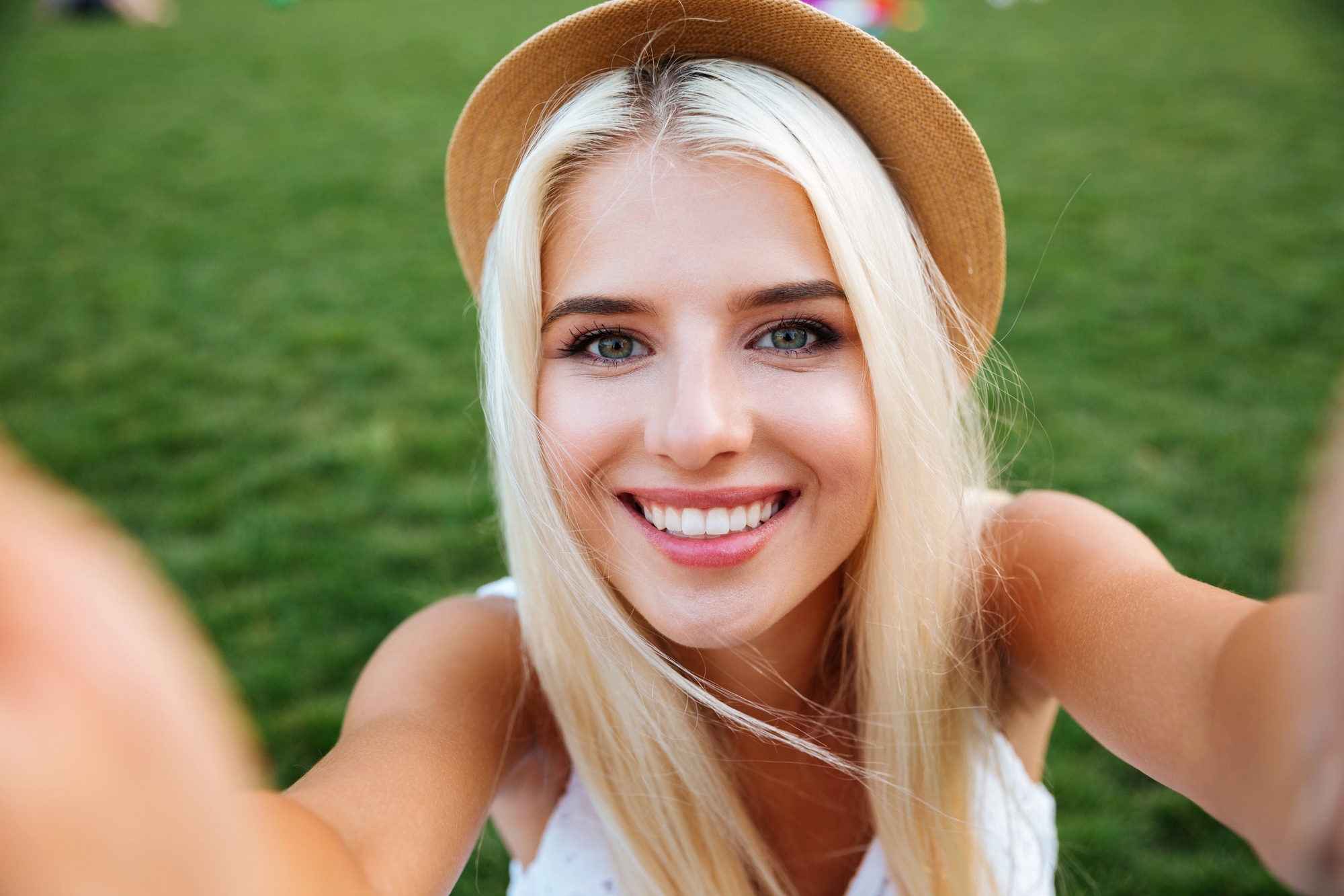 A person with long blonde hair wearing a straw hat takes a selfie outdoors. They are smiling widely, with a grassy background. The sun is shining, creating a bright and warm atmosphere.