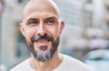 A smiling bald man with a salt-and-pepper beard stands outdoors in soft focus. He wears a light-colored shirt and appears to be enjoying the day. The background is a blurred urban setting, giving a casual, relaxed atmosphere.