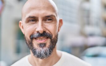 A smiling bald man with a salt-and-pepper beard stands outdoors in soft focus. He wears a light-colored shirt and appears to be enjoying the day. The background is a blurred urban setting, giving a casual, relaxed atmosphere.