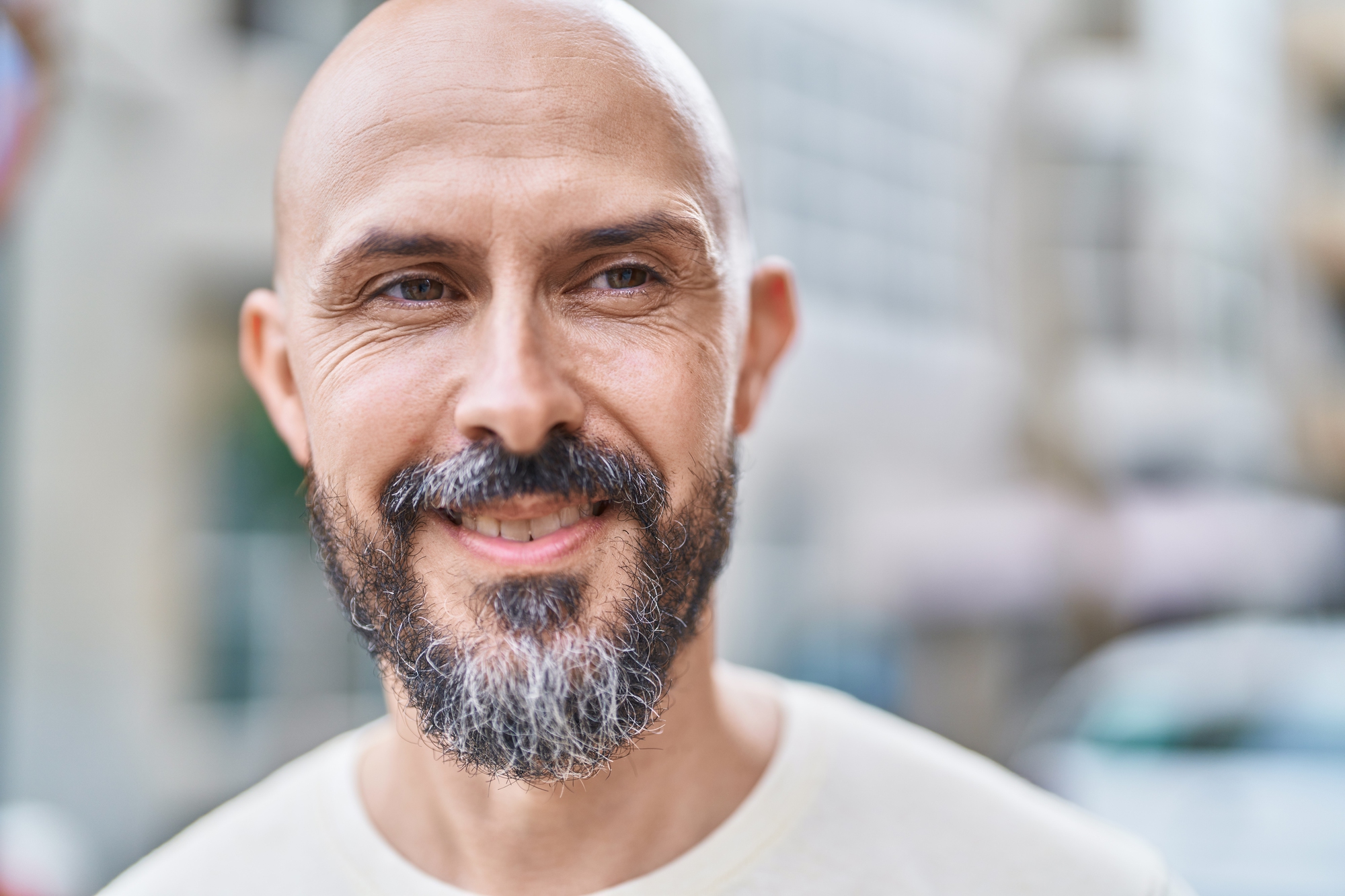 A smiling bald man with a salt-and-pepper beard stands outdoors in soft focus. He wears a light-colored shirt and appears to be enjoying the day. The background is a blurred urban setting, giving a casual, relaxed atmosphere.