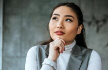 A thoughtful person with long hair and red lipstick gazes upward. They are wearing a gray sleeveless jacket over a white turtleneck and resting their chin on their hand. The background is a blurred gray wall.