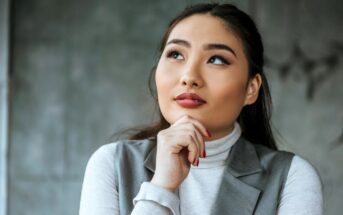 A thoughtful person with long hair and red lipstick gazes upward. They are wearing a gray sleeveless jacket over a white turtleneck and resting their chin on their hand. The background is a blurred gray wall.