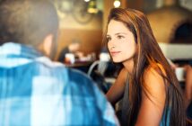 A woman with long brown hair sits across from a man in a cozy café. She is looking at him attentively, with blurred people and warm, rustic decor in the background.