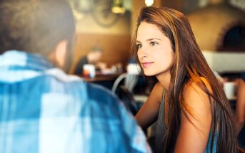 A woman with long brown hair sits across from a man in a cozy café. She is looking at him attentively, with blurred people and warm, rustic decor in the background.