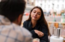 A woman with long brown hair sits at an outdoor table, gazing thoughtfully at a person sitting across from her. She wears a dark jacket, and a bowl of food and glass bottles are on the table. The background shows a blurred cityscape.