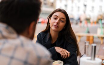 A woman with long brown hair sits at an outdoor table, gazing thoughtfully at a person sitting across from her. She wears a dark jacket, and a bowl of food and glass bottles are on the table. The background shows a blurred cityscape.