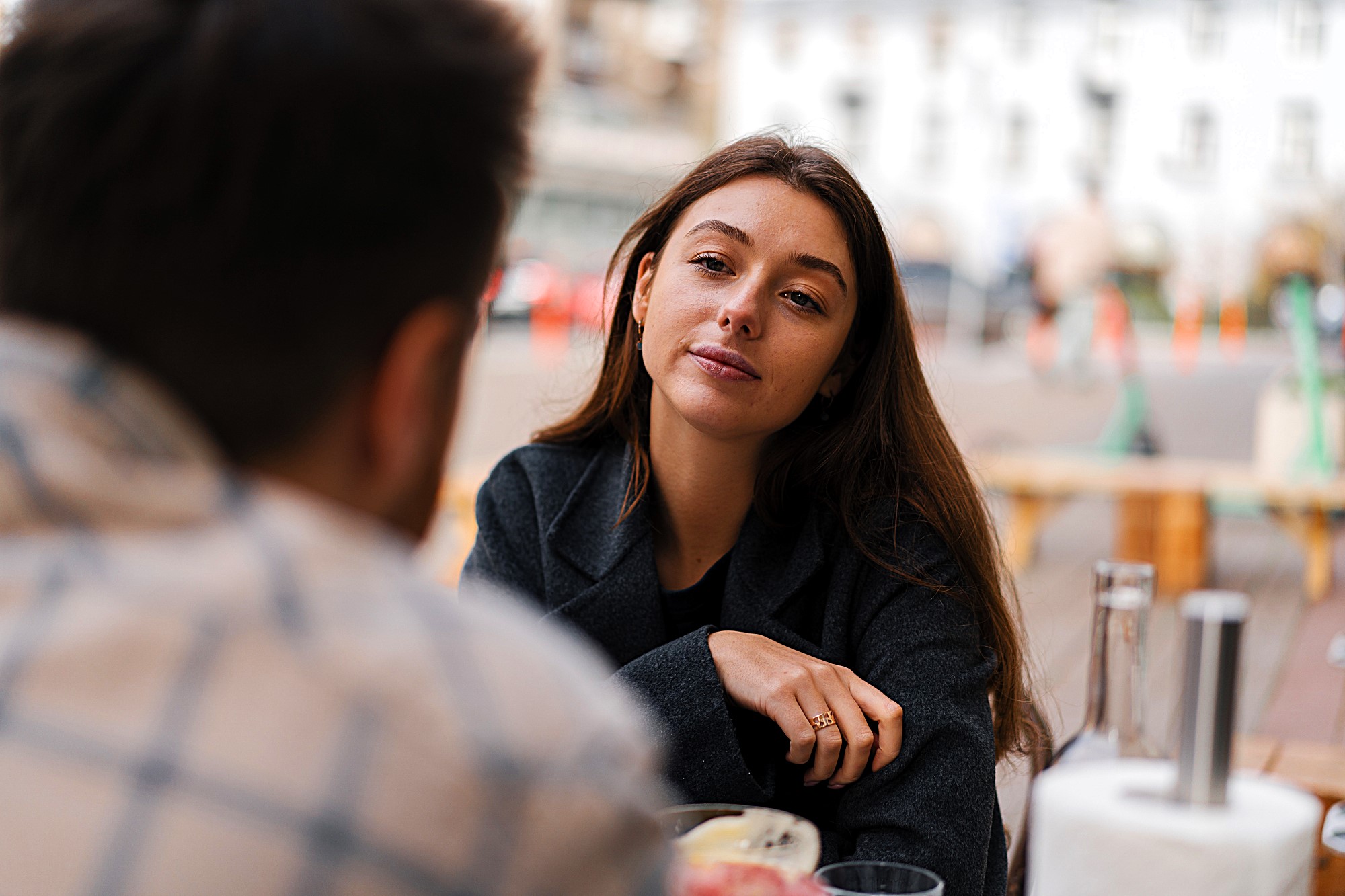 A woman with long brown hair sits at an outdoor table, gazing thoughtfully at a person sitting across from her. She wears a dark jacket, and a bowl of food and glass bottles are on the table. The background shows a blurred cityscape.