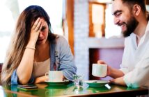 A woman looks distressed with her hand on her forehead, while a man laughs beside her. Both are seated at a table with coffee cups in a cozy café setting.