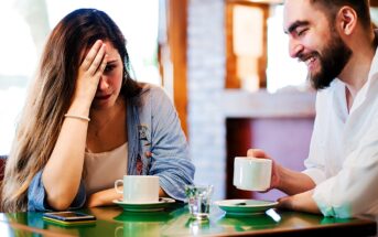 A woman looks distressed with her hand on her forehead, while a man laughs beside her. Both are seated at a table with coffee cups in a cozy café setting.