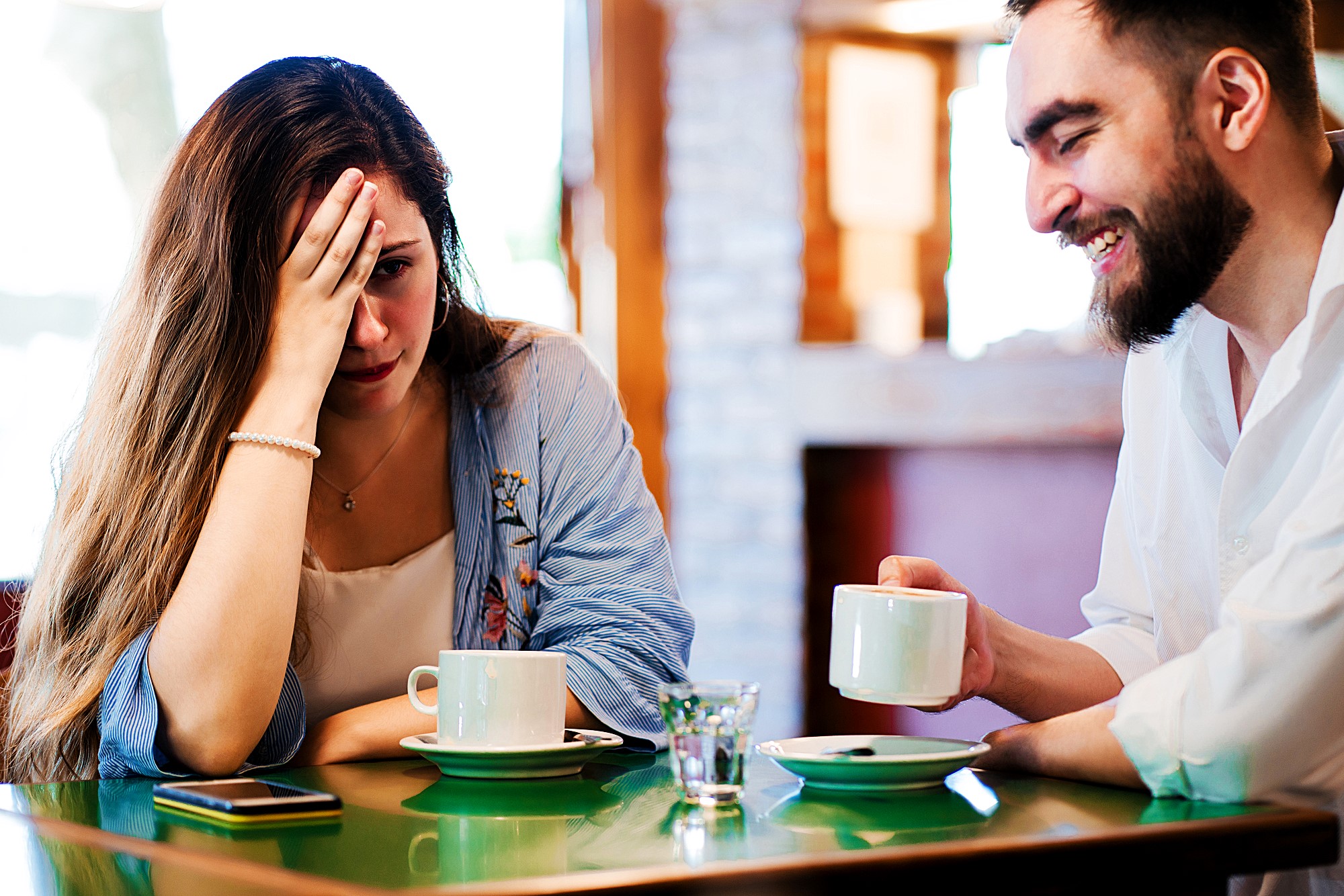 A woman looks distressed with her hand on her forehead, while a man laughs beside her. Both are seated at a table with coffee cups in a cozy café setting.