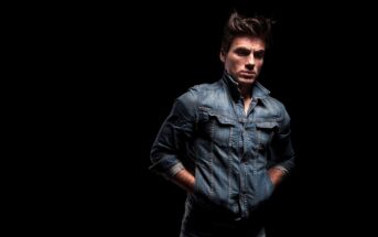 A man wearing a denim jacket poses against a dark background. His hands are in his pockets, and the lighting highlights his face and the texture of the denim.