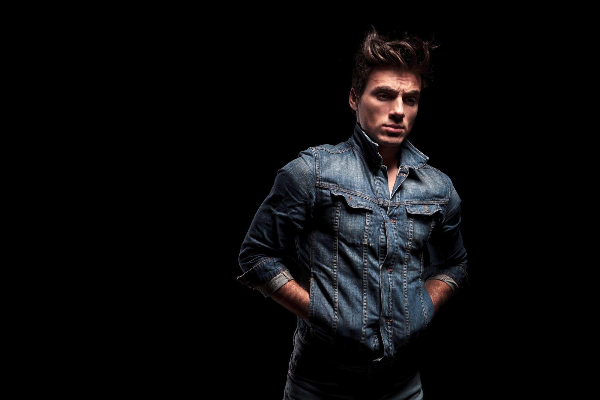 A man wearing a denim jacket poses against a dark background. His hands are in his pockets, and the lighting highlights his face and the texture of the denim.