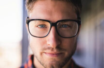 A person with short hair and a beard wears black-framed glasses, looking directly at the camera with a neutral expression. The background is blurred.