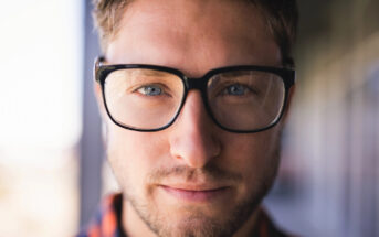 A person with short hair and a beard wears black-framed glasses, looking directly at the camera with a neutral expression. The background is blurred.