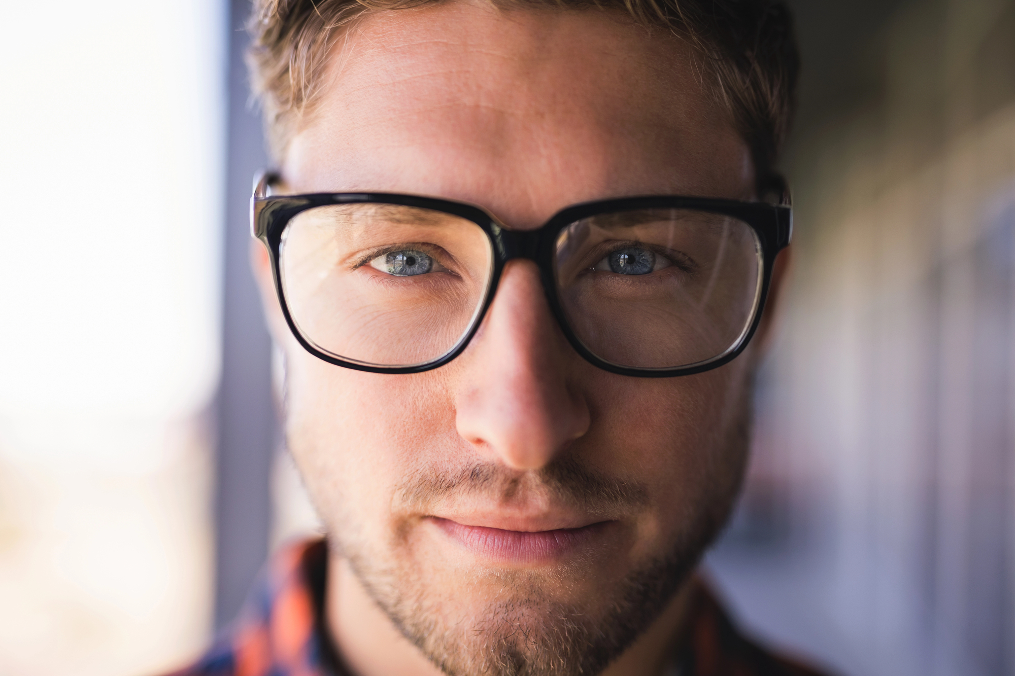 A person with short hair and a beard wears black-framed glasses, looking directly at the camera with a neutral expression. The background is blurred.