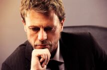 A man with a thoughtful expression rests his chin on his hand. He is wearing a dark pinstripe suit and is seated against a neutral-colored background. The lighting highlights his pensive demeanor.