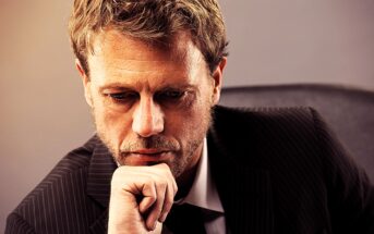 A man with a thoughtful expression rests his chin on his hand. He is wearing a dark pinstripe suit and is seated against a neutral-colored background. The lighting highlights his pensive demeanor.