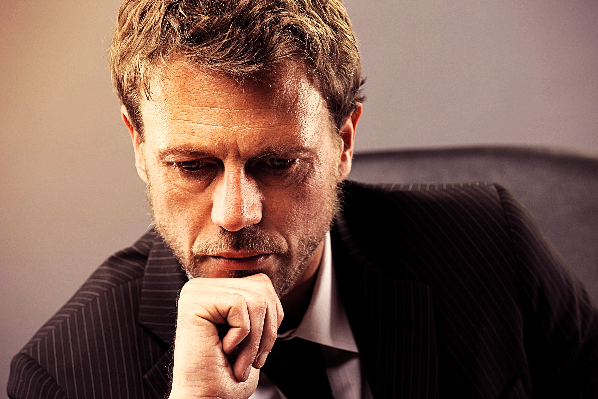 A man with a thoughtful expression rests his chin on his hand. He is wearing a dark pinstripe suit and is seated against a neutral-colored background. The lighting highlights his pensive demeanor.