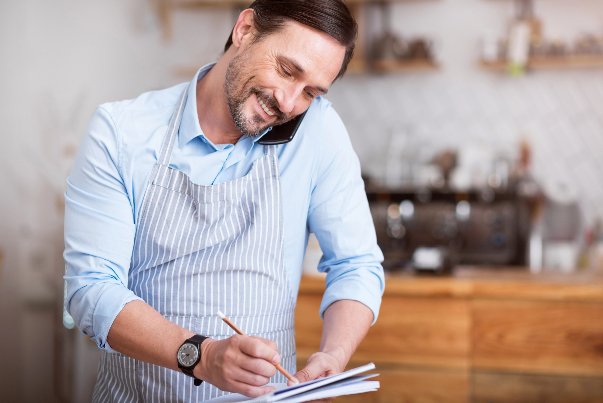 A man wearing an apron is smiling while talking on a phone pressed between his ear and shoulder. He is writing on a notepad. The background shows a kitchen setting with wooden cabinets and a coffee machine.