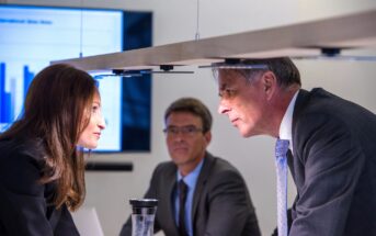 Three business professionals in a meeting room, with two engaged in a focused discussion across a conference table. A screen displaying a bar graph is in the background. One person is observing attentively.