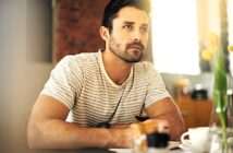 A man with short dark hair and a beard is sitting indoors, wearing a striped shirt. He gazes thoughtfully to the side, resting his arms on a table with a cup and a pot of colored pencils nearby. Tulips in a vase are partially visible.